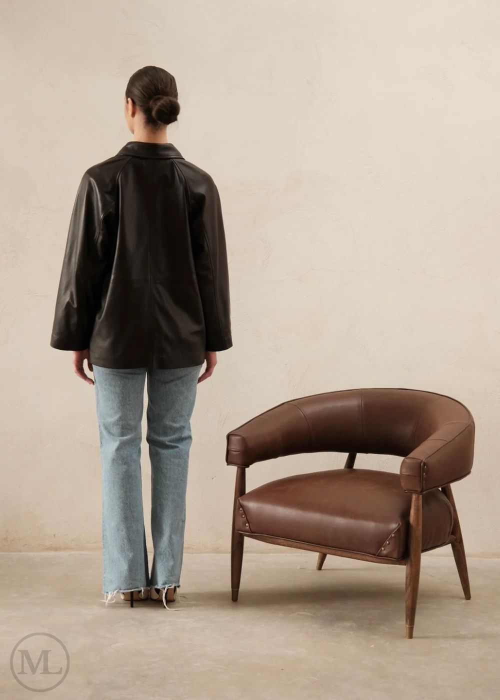Women wearing a dark brown leather trucker jacket and light blue jeans standing next to a brown leather chair against a beige wall.
