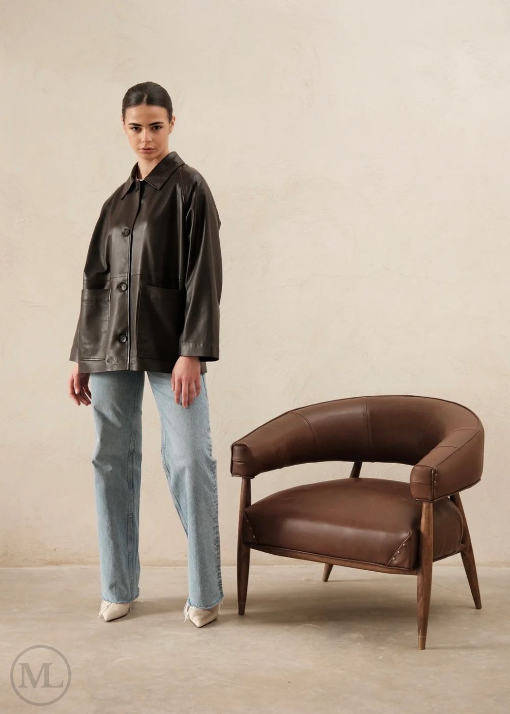 Woman in a dark brown leather trucker jacket and light blue jeans standing next to a brown leather chair against a beige wall.