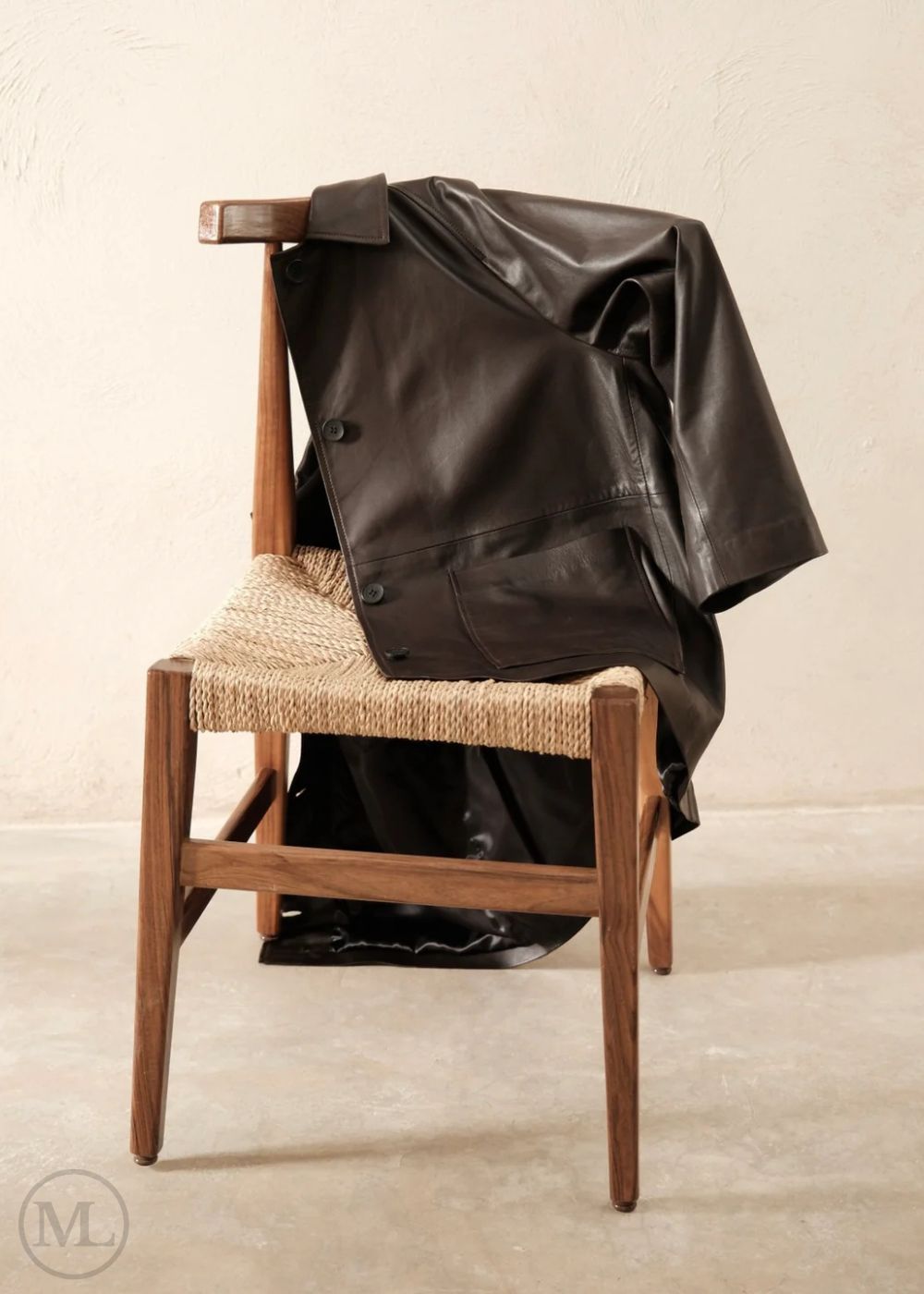 Dark brown leather trucker jacket draped over a wooden chair with a woven seat on a plain background
