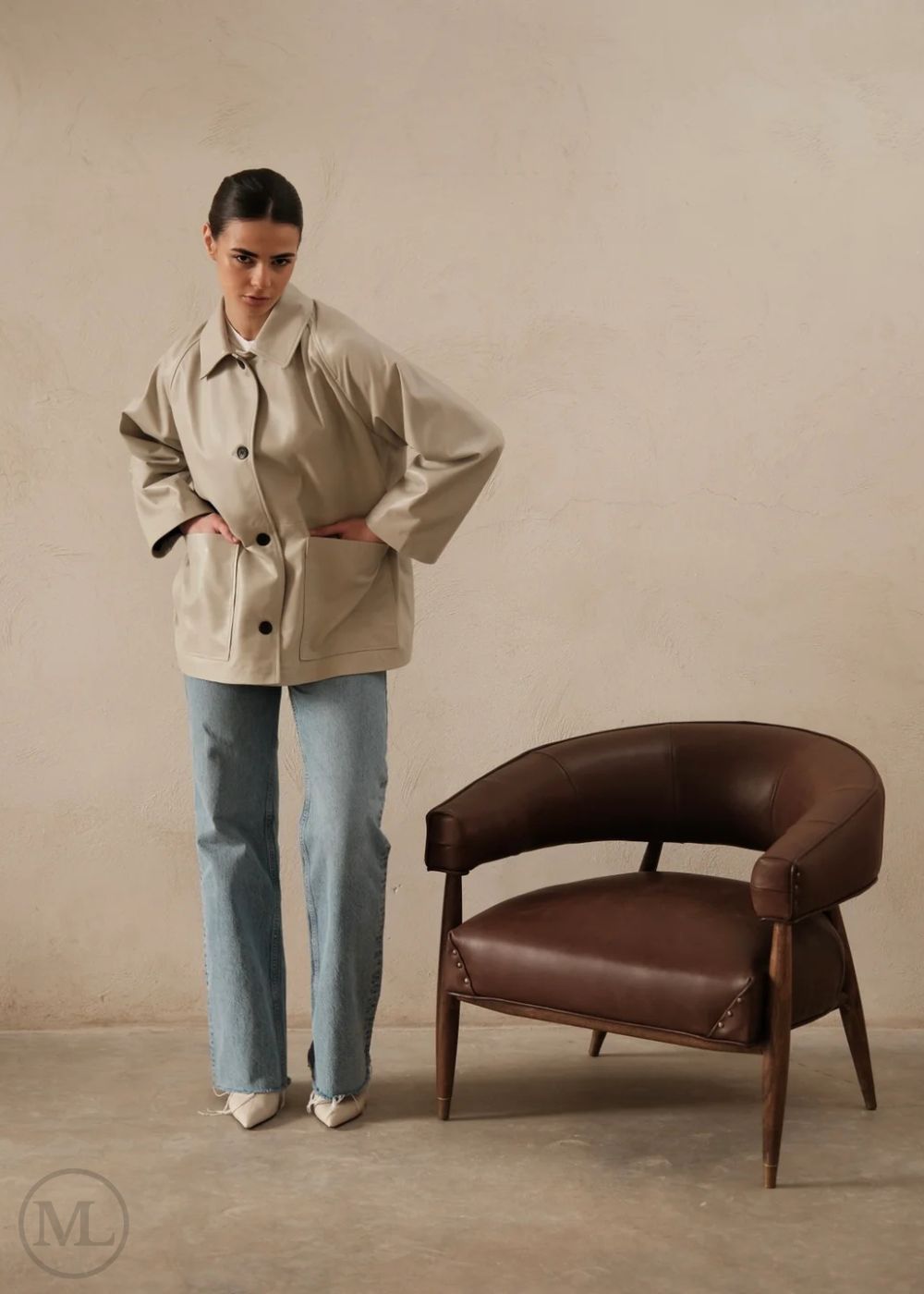 Women wearing a lvory leather blazer jacket and light blue jeans standing next to a brown leather chair against a beige wall.