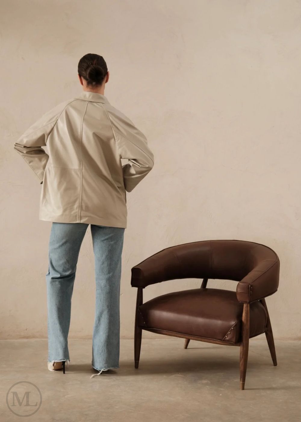 Women wearing a lvory leather blazer jacket and light blue jeans standing next to a brown leather chair against a beige wall.