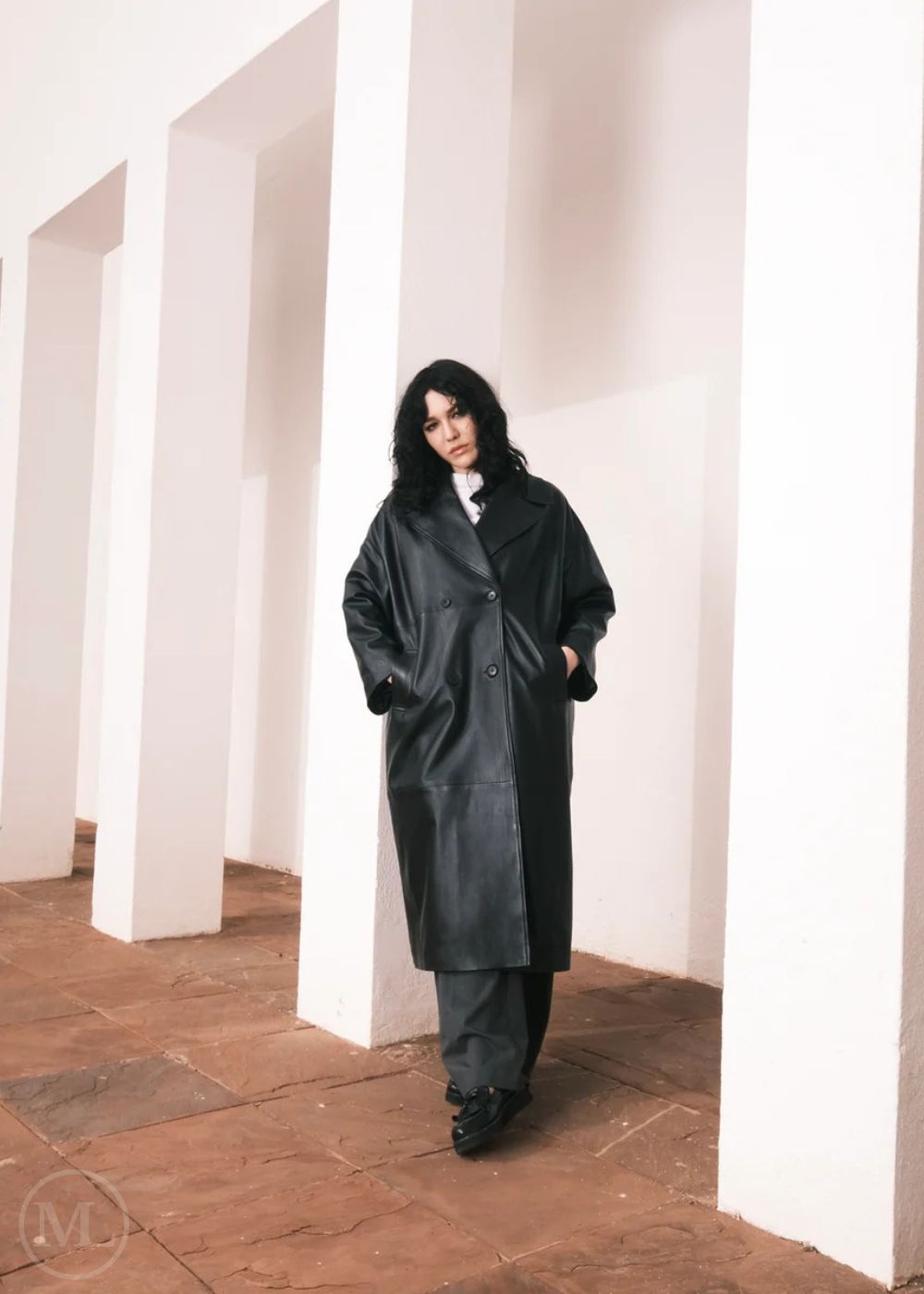 Femmes portant un long trench-coat en cuir, debout dans un décor minimaliste avec des colonnes blanches.