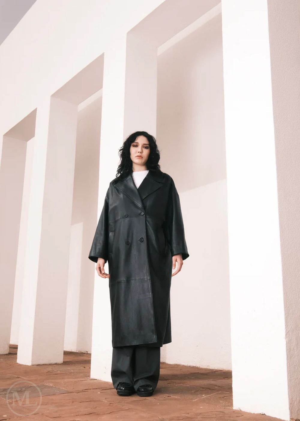 Femmes portant un long trench-coat en cuir, debout dans un décor minimaliste avec des colonnes blanches.