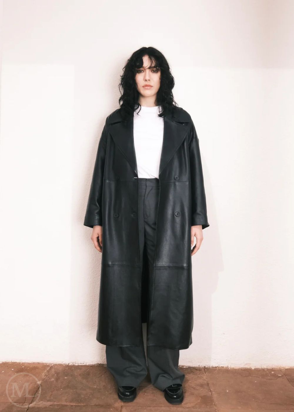 Femmes portant un long trench-coat en cuir, debout dans un décor minimaliste avec des colonnes blanches.