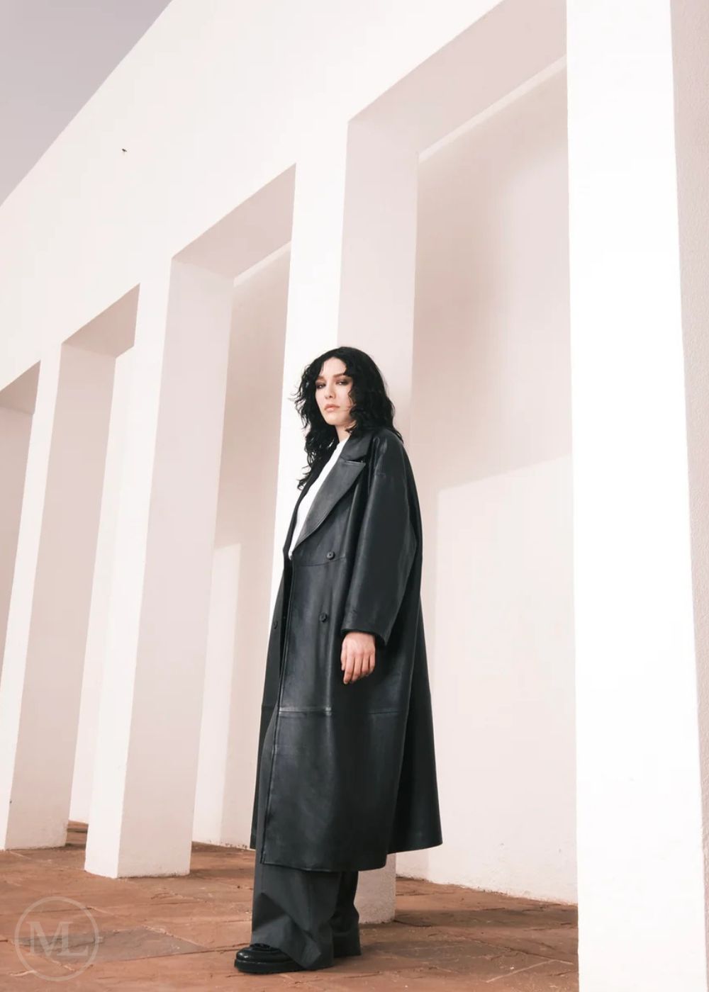 Femmes portant un long trench-coat en cuir, debout dans un décor minimaliste avec des colonnes blanches.