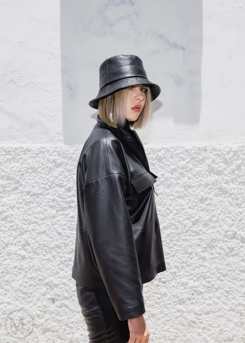 Women wearing a black leather jacket and bucket hat against a textured white wall