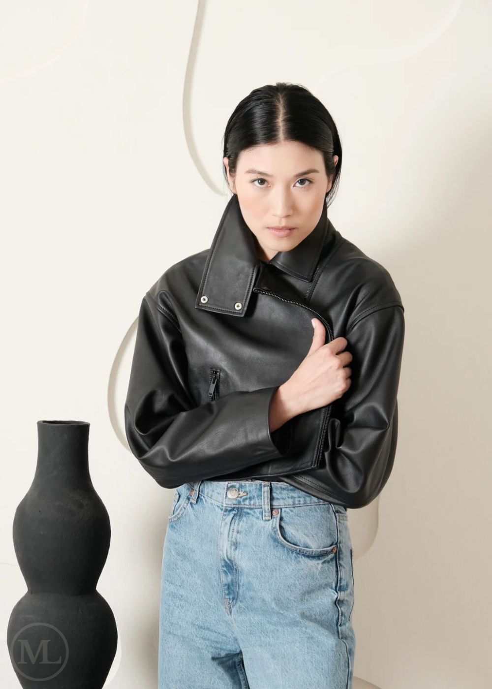 Women wearing a Cropped black leather Biker jacket and light blue jeans standing next to a black vase on a white background
