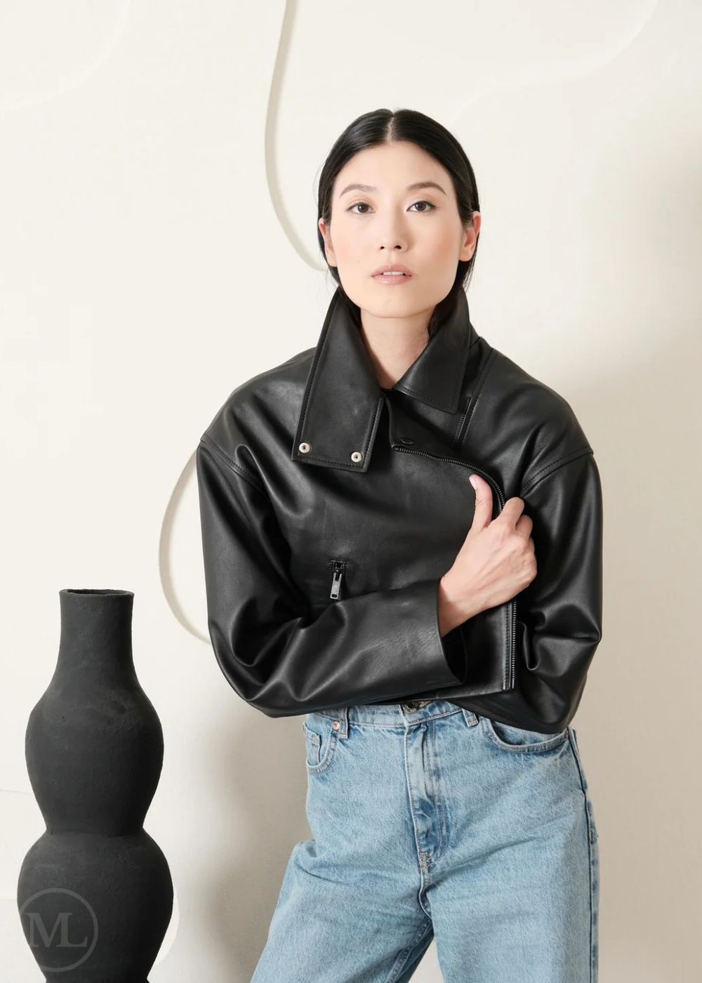 Women wearing a Cropped black leather Biker jacket and light blue jeans standing next to a black vase on a white background