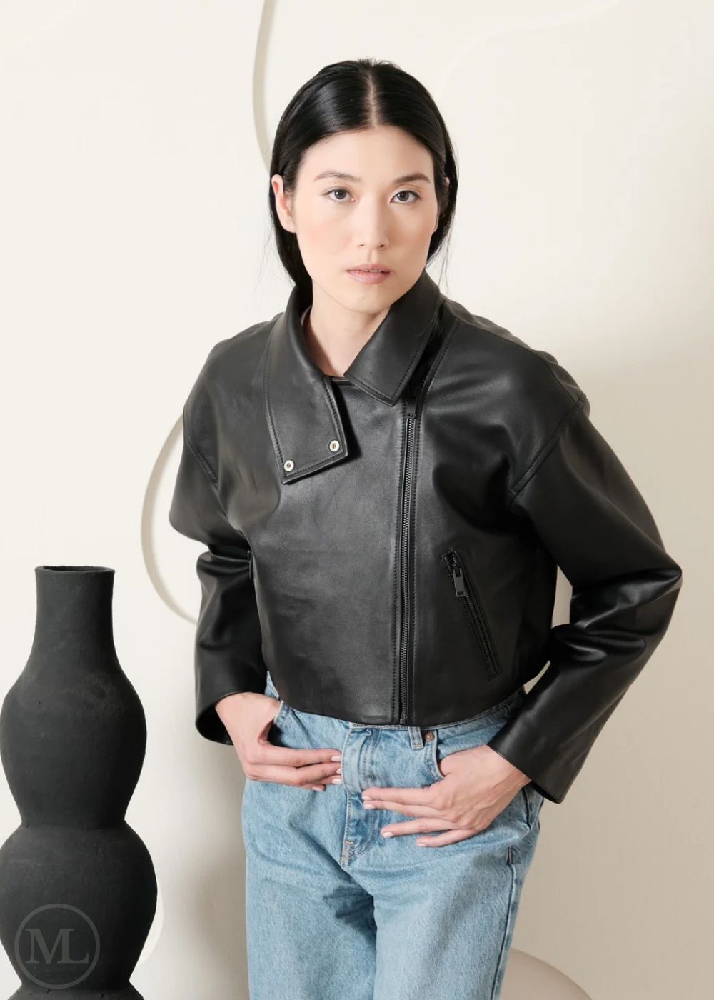 Women wearing a Cropped black leather Biker jacket and light blue jeans standing next to a black vase on a white background