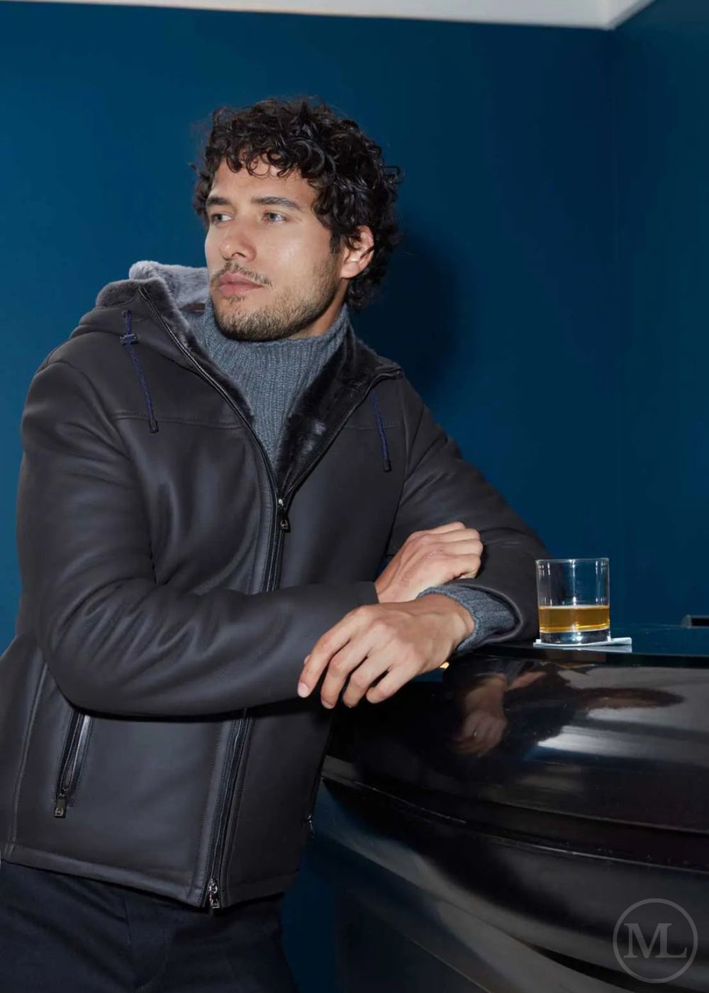 Man in a dark grey hooded shearling jacket leaning against a car with a drink in a glass, blue wall background