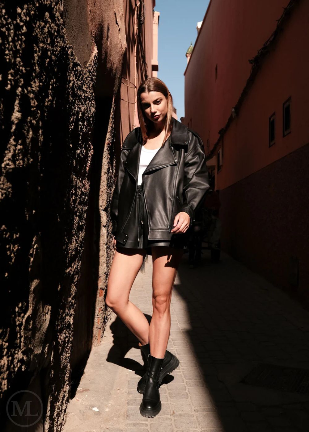 Woman in a leather biker jacket and black boots standing in a narrow alleyway.