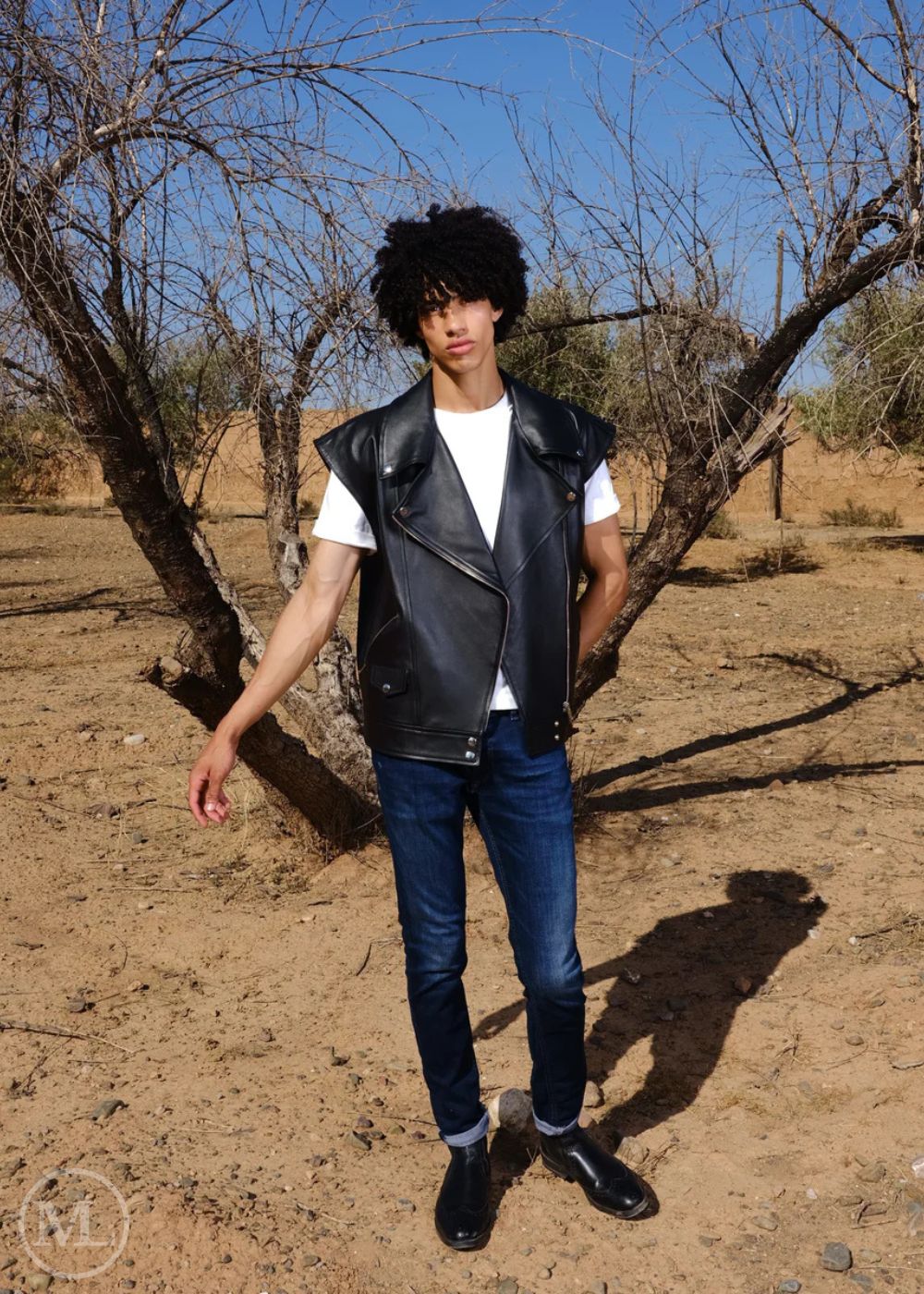 Women wearing a black leather vest, white shirt, blue jeans, and black shoes standing in a dry, barren landscape.