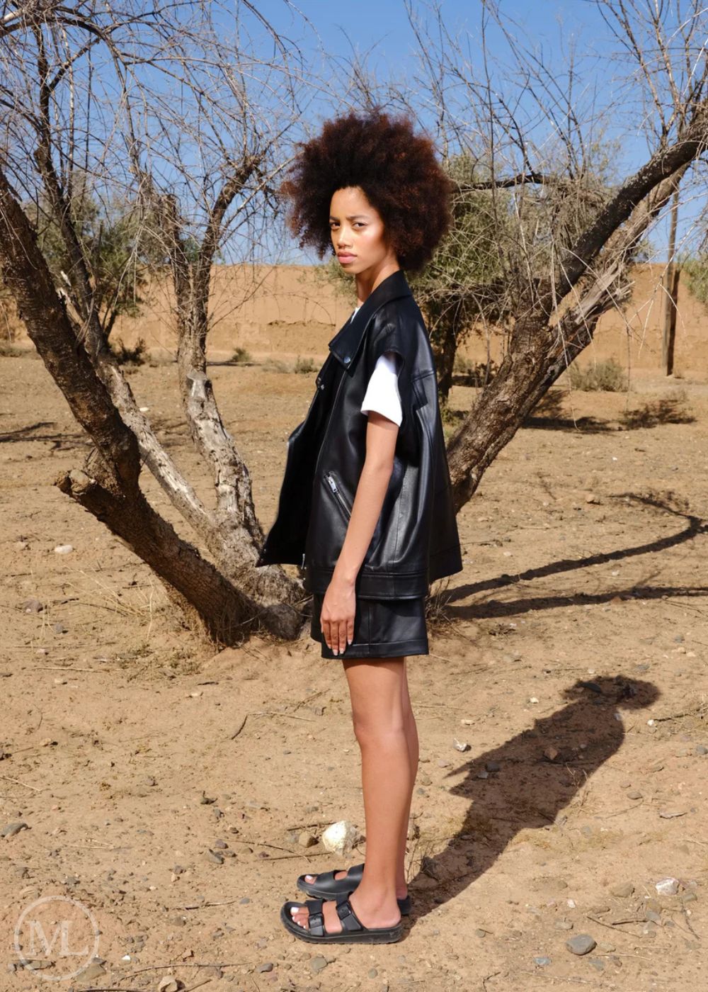 Women wearing a black vest and shorts standing in a desert-like environment with trees and a clear sky.