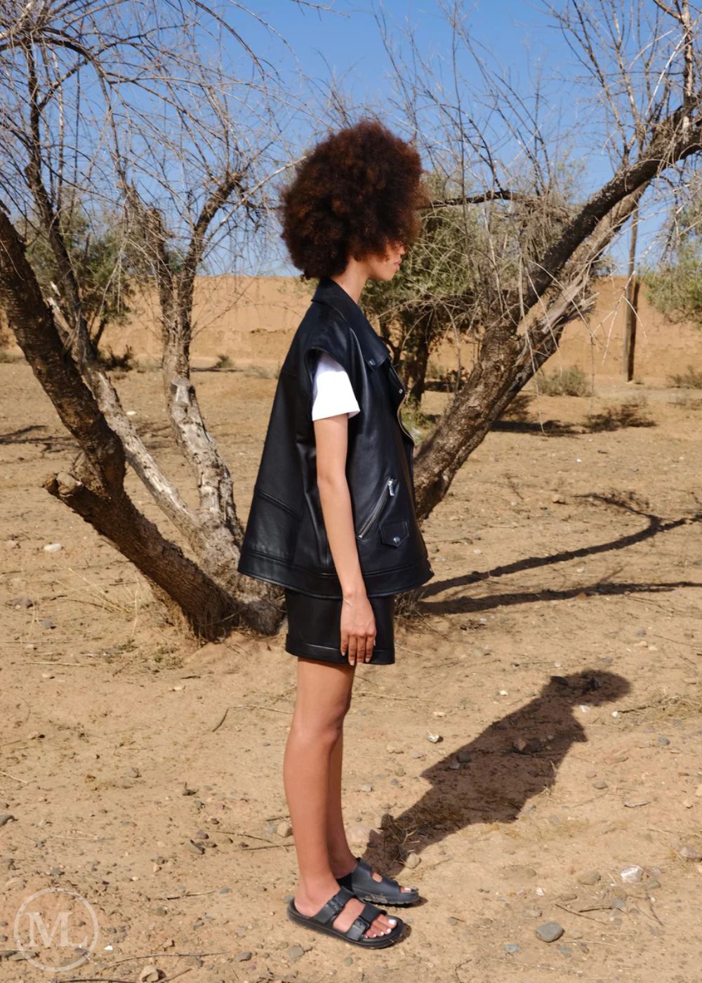 Women wearing a black leather vest and sandals standing under a tree in a desert-like setting.