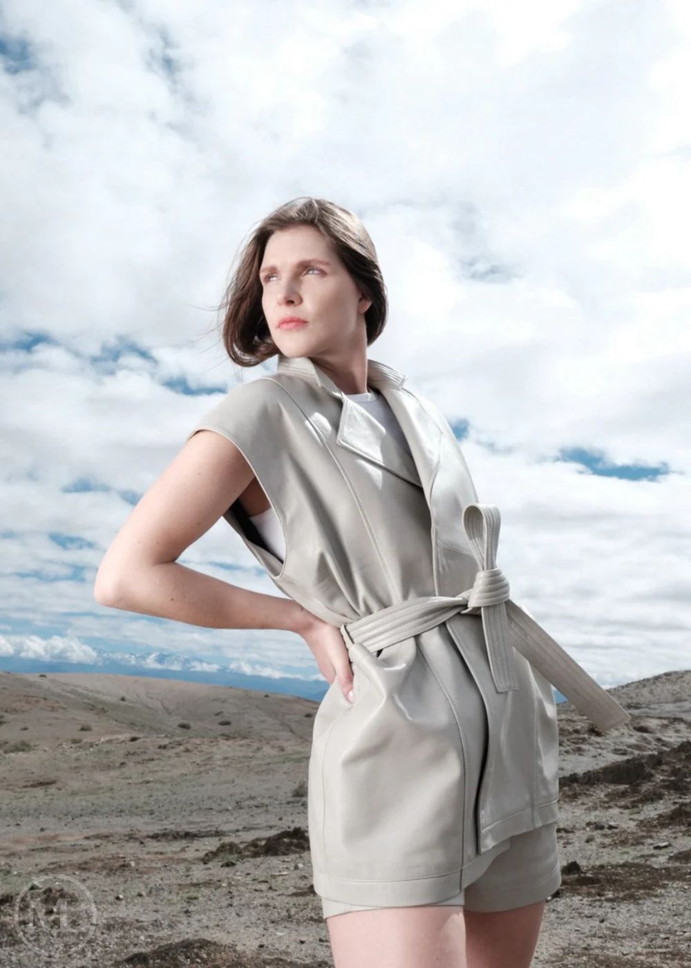 Woman in a lvory outfit standing against a scenic background with clouds and mountains.