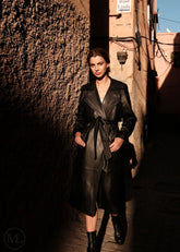 Woman in a black leather trench coat standing in a narrow alleyway.