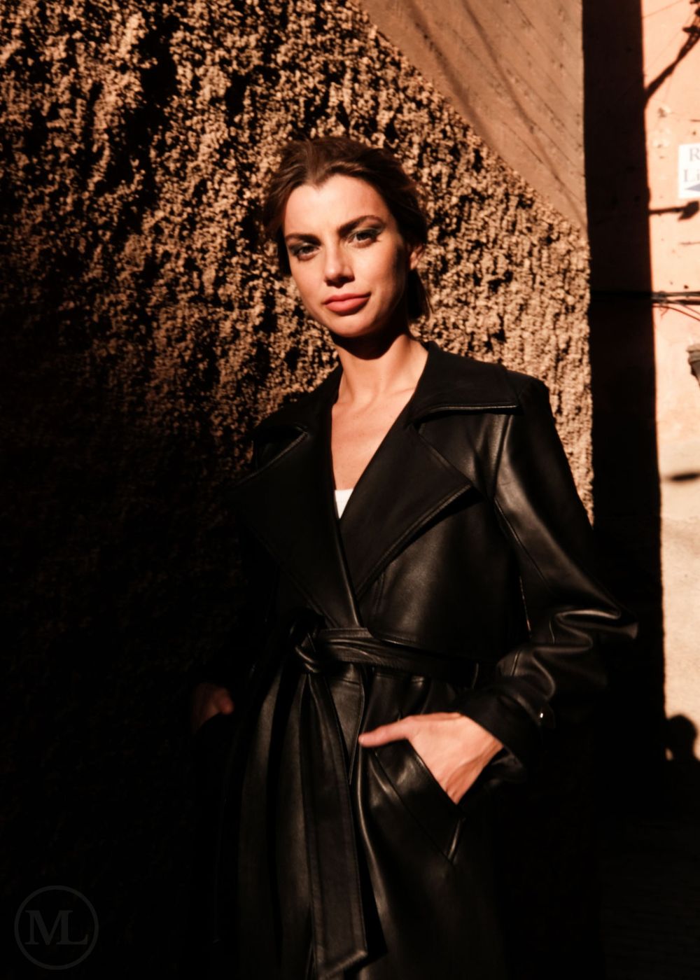 Woman wearing a black leather trench coat standing against a textured wall.