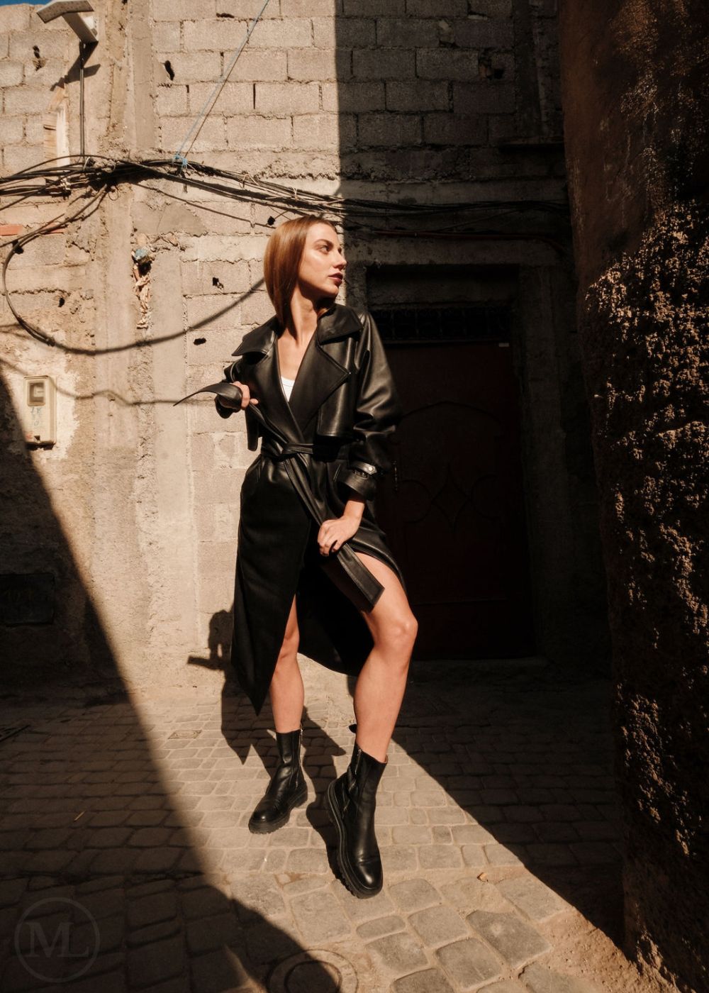 Woman in a black leather trench coat standing in a sunlit alleyway.