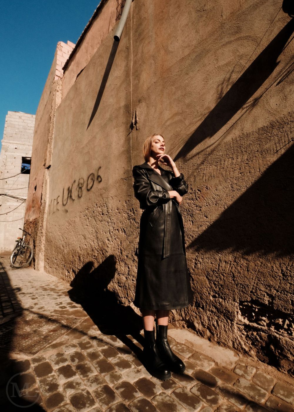Woman in a black leather trench coat standing against a textured wall with graffiti.