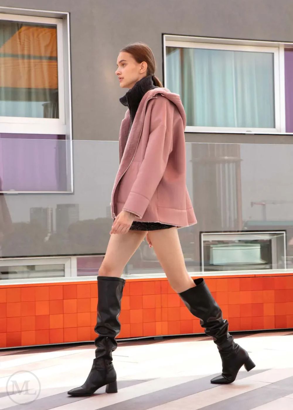 Woman in a fuschi leather hooded jacket and black knee-high boots walking past a colorful train.
