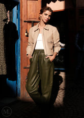 Woman in beige jacket and green pants standing in a dimly lit room with wooden door.