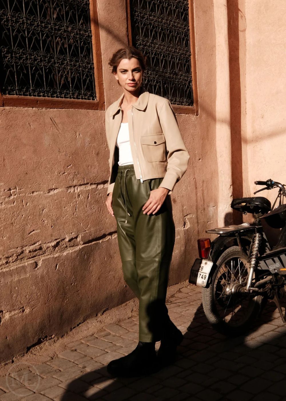 Woman in beige leather cropped jacket and green pants standing next to a motorcycle against a textured wall.