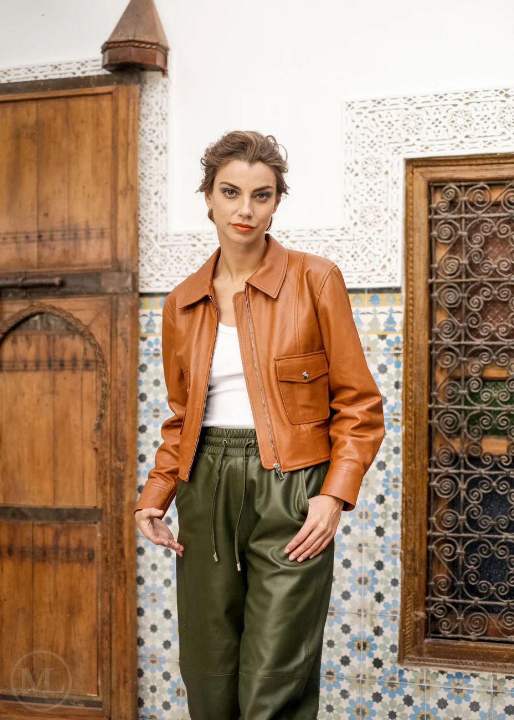 Woman wearing a brown leather jacket and green pants standing in front of decorative doors and wall.