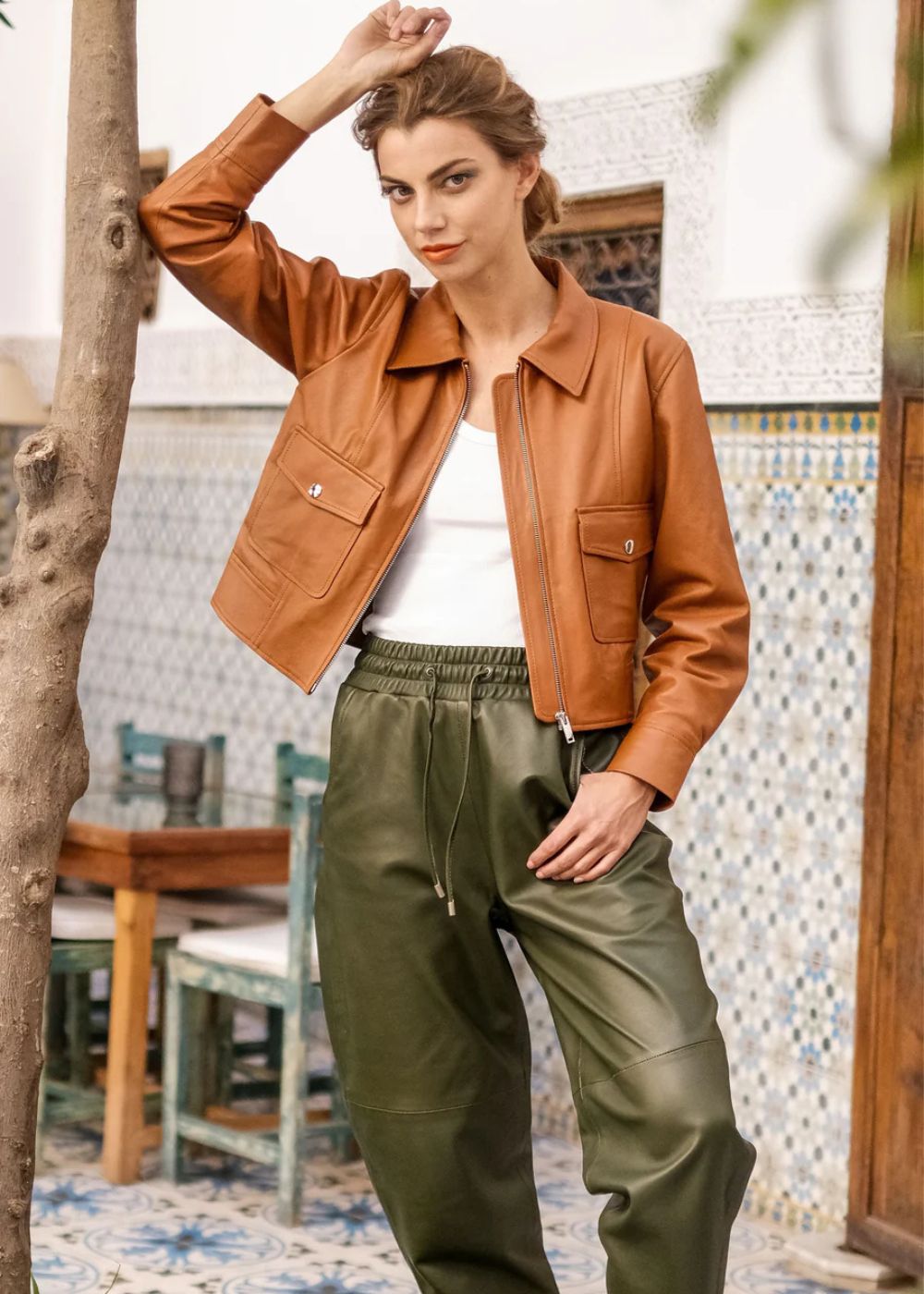 Woman wearing a brown leather jacket and green pants standing in an outdoor setting with tiled walls.
