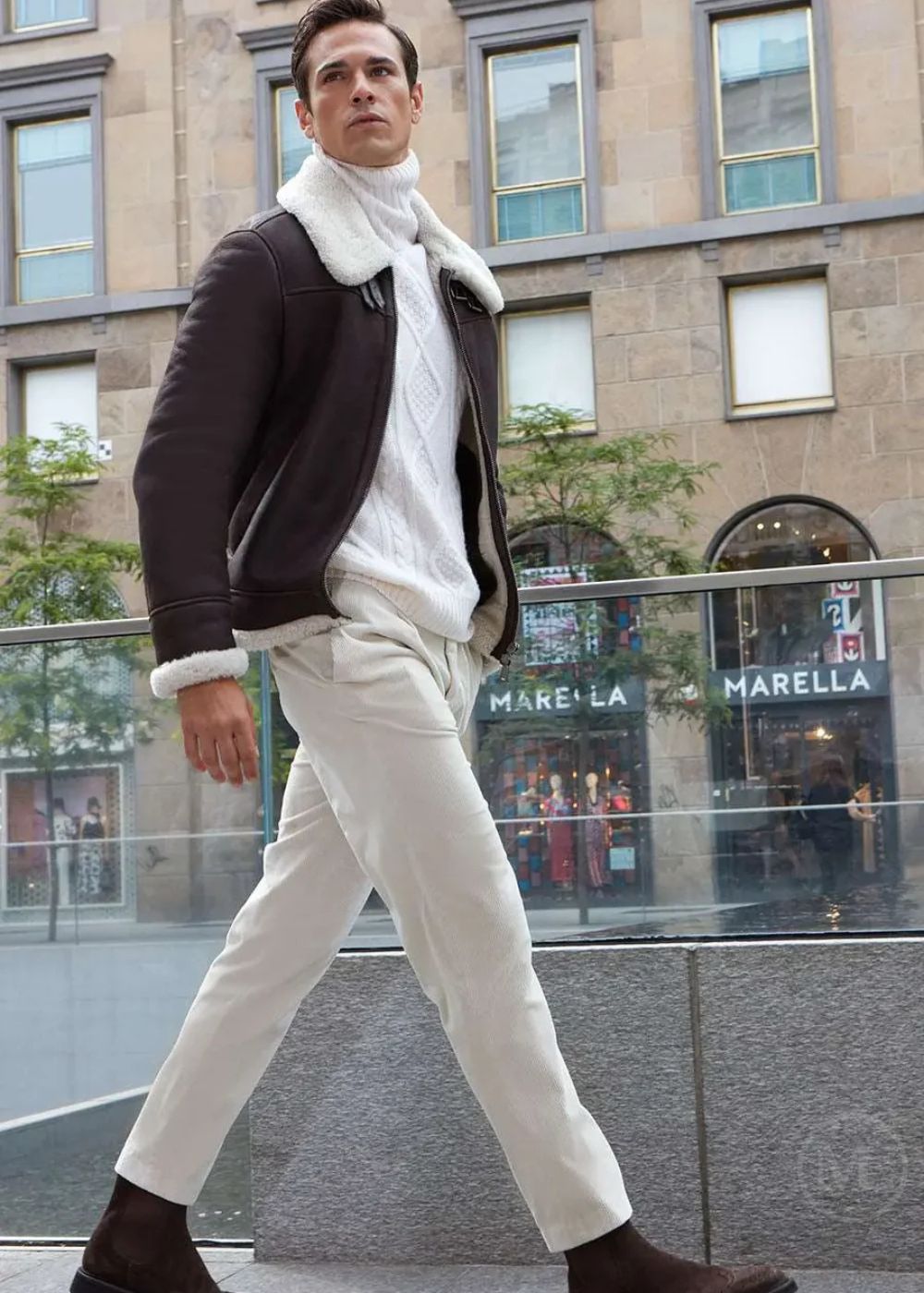 Man walking on a city street wearing a brown shearling aviator jacket with white fur trim, white sweater, and white pants.