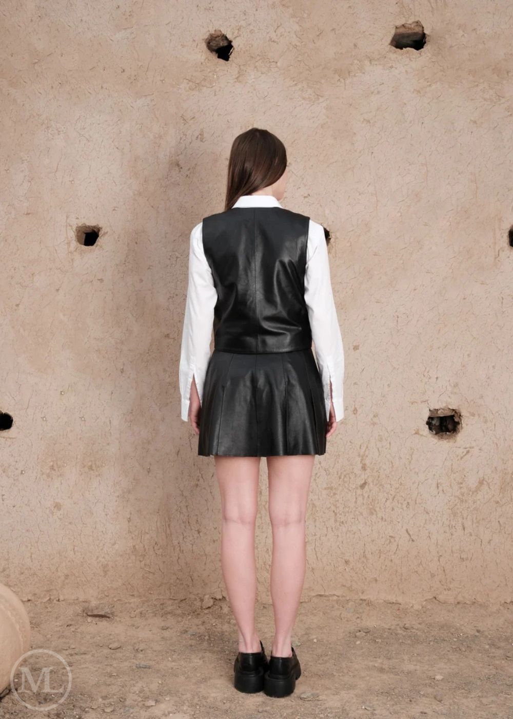 Women wearing a black leather vest and skirt against a textured beige wall.