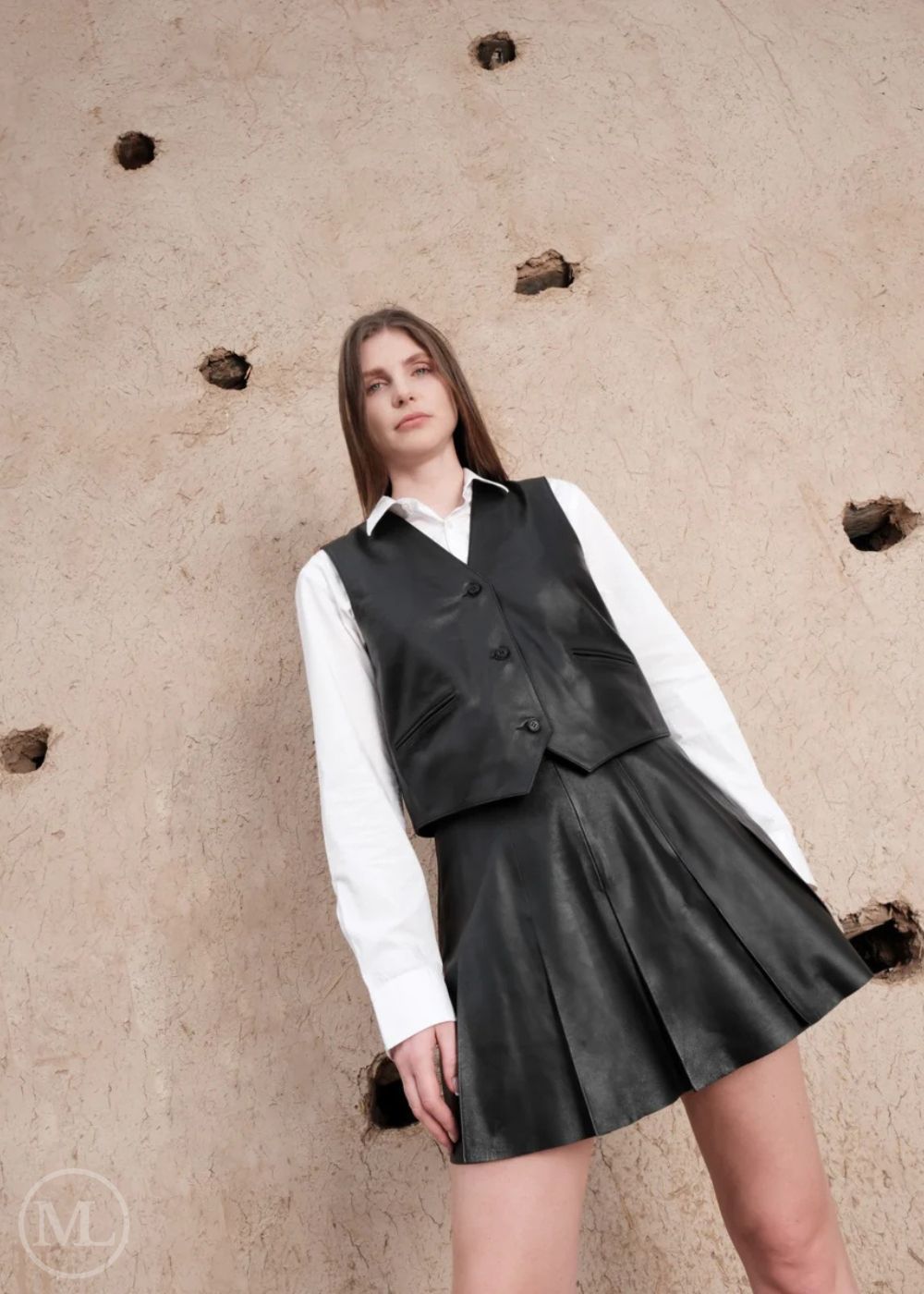 Woman wearing a black vest and skirt over a white shirt against a textured beige wall.
