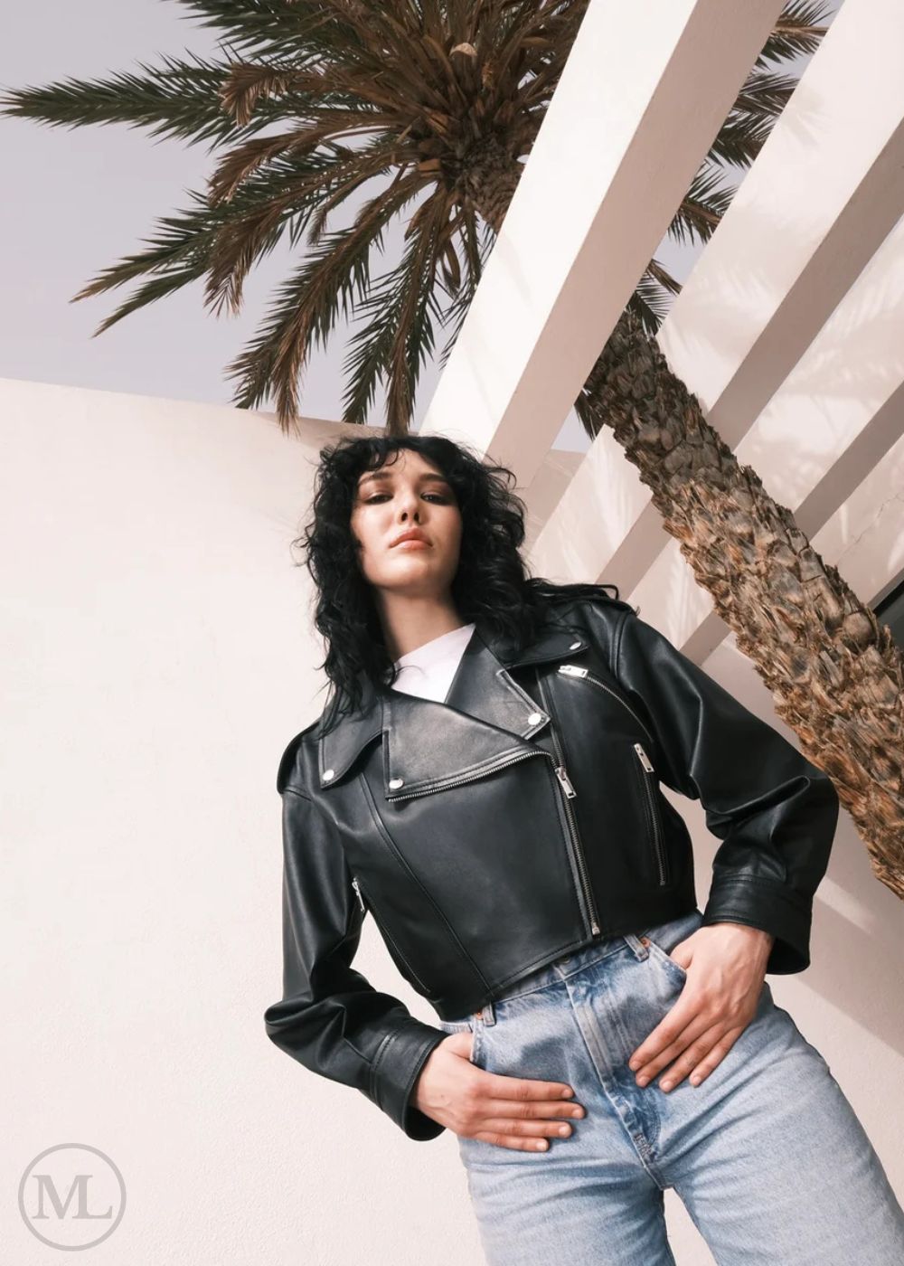 Women wearing a Cropped black leather Biker jacket and light blue jeans standing against a neutral wall with palm trees.