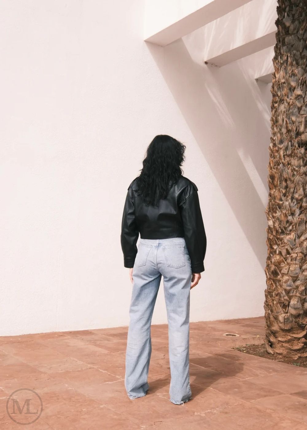 Women wearing a Cropped black leather Biker jacket and light blue jeans standing against a neutral wall with palm trees.