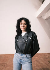 Women wearing a Cropped black leather Biker jacket and light blue jeans standing against a neutral wall with palm trees.