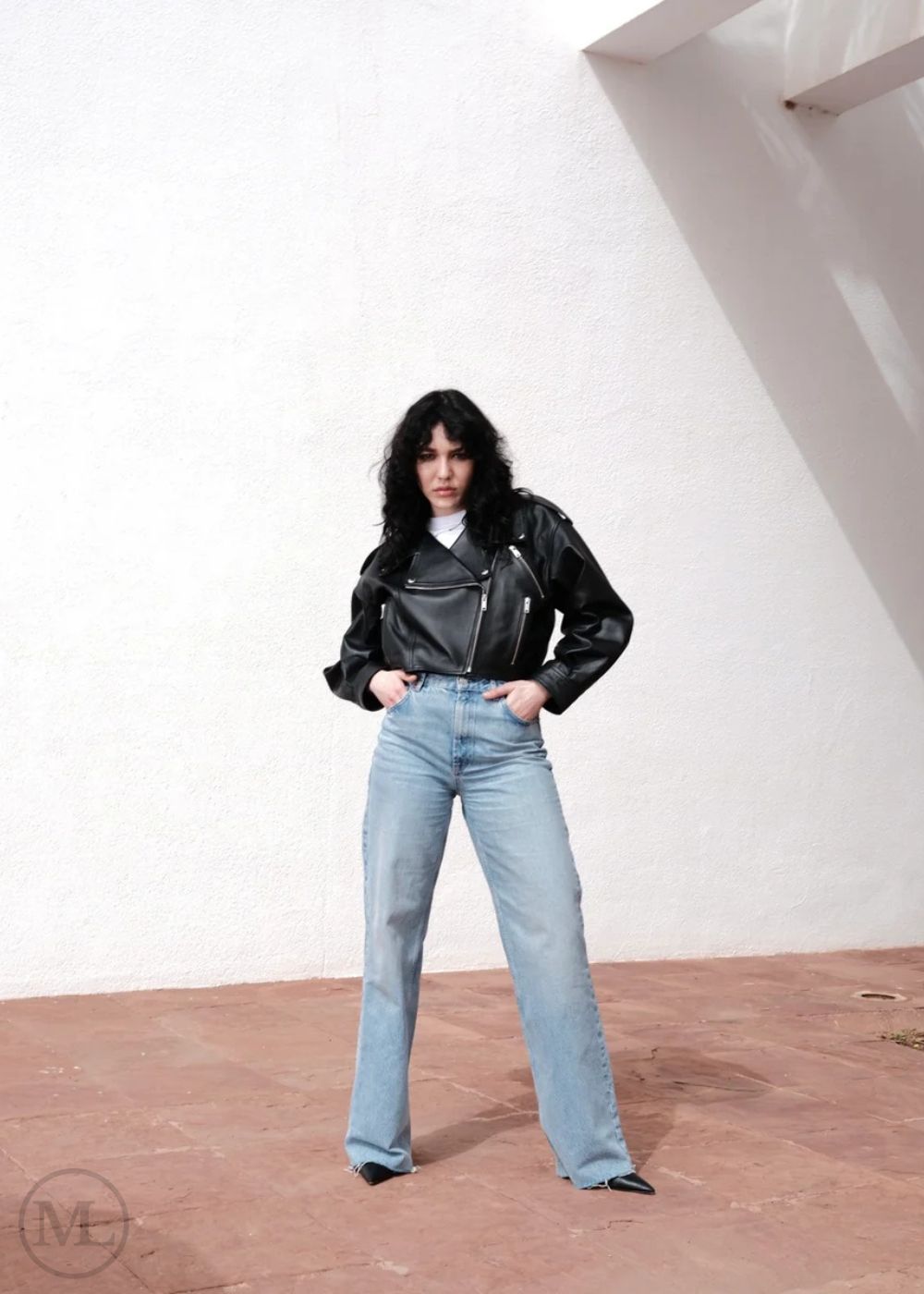 Women wearing a Cropped black leather Biker jacket and light blue jeans standing against a neutral wall with palm trees.