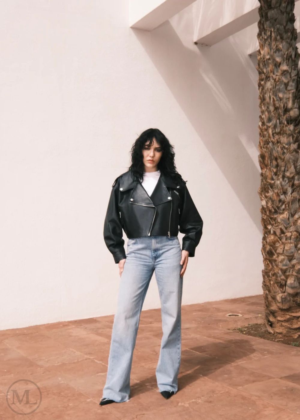 Women wearing a Cropped black leather Biker jacket and light blue jeans standing against a neutral wall with palm trees.