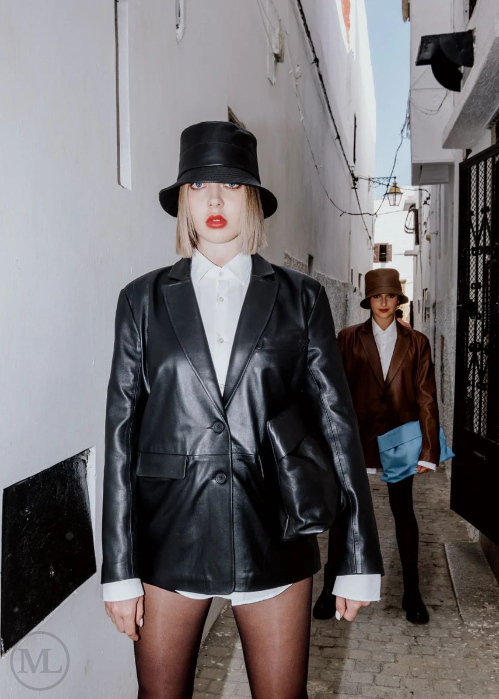 Woman wearing a black blazer Coat, white shirt, and hat standing against a beige wall.