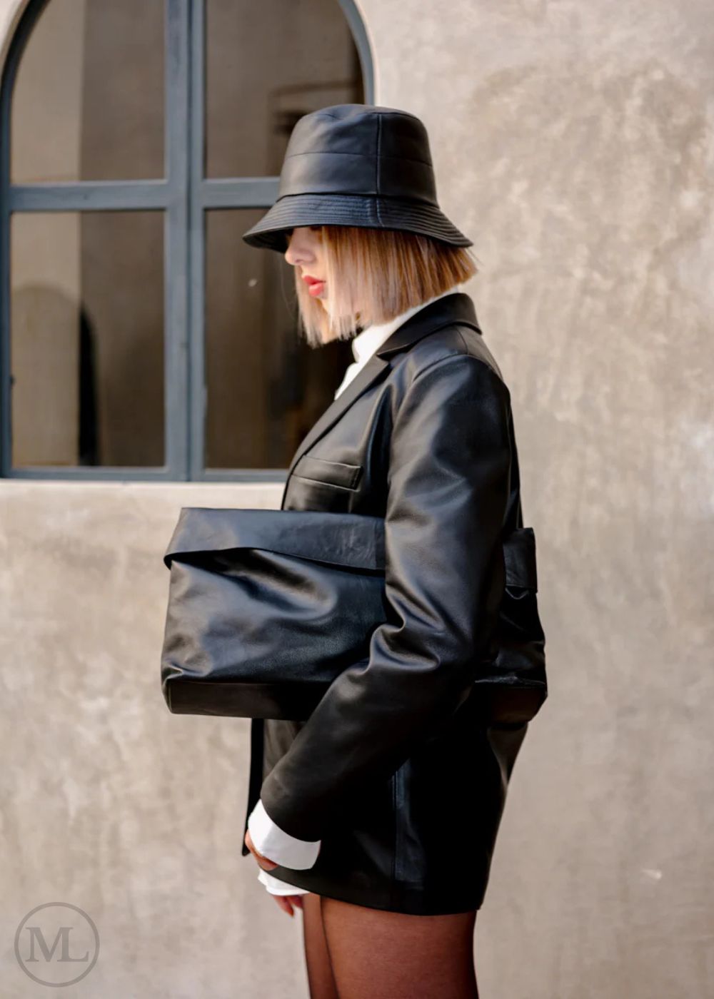 Woman wearing a black blazer Coat, white shirt, and hat standing against a beige wall.