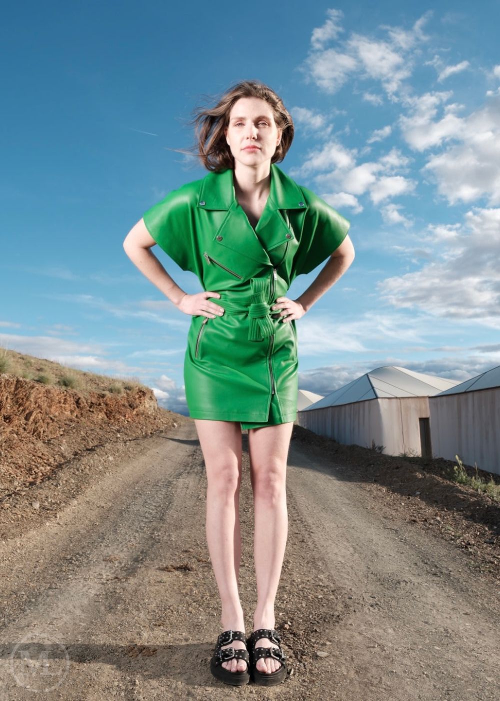 Woman in a green leather dress standing on a dirt road with a blue sky and clouds in the background