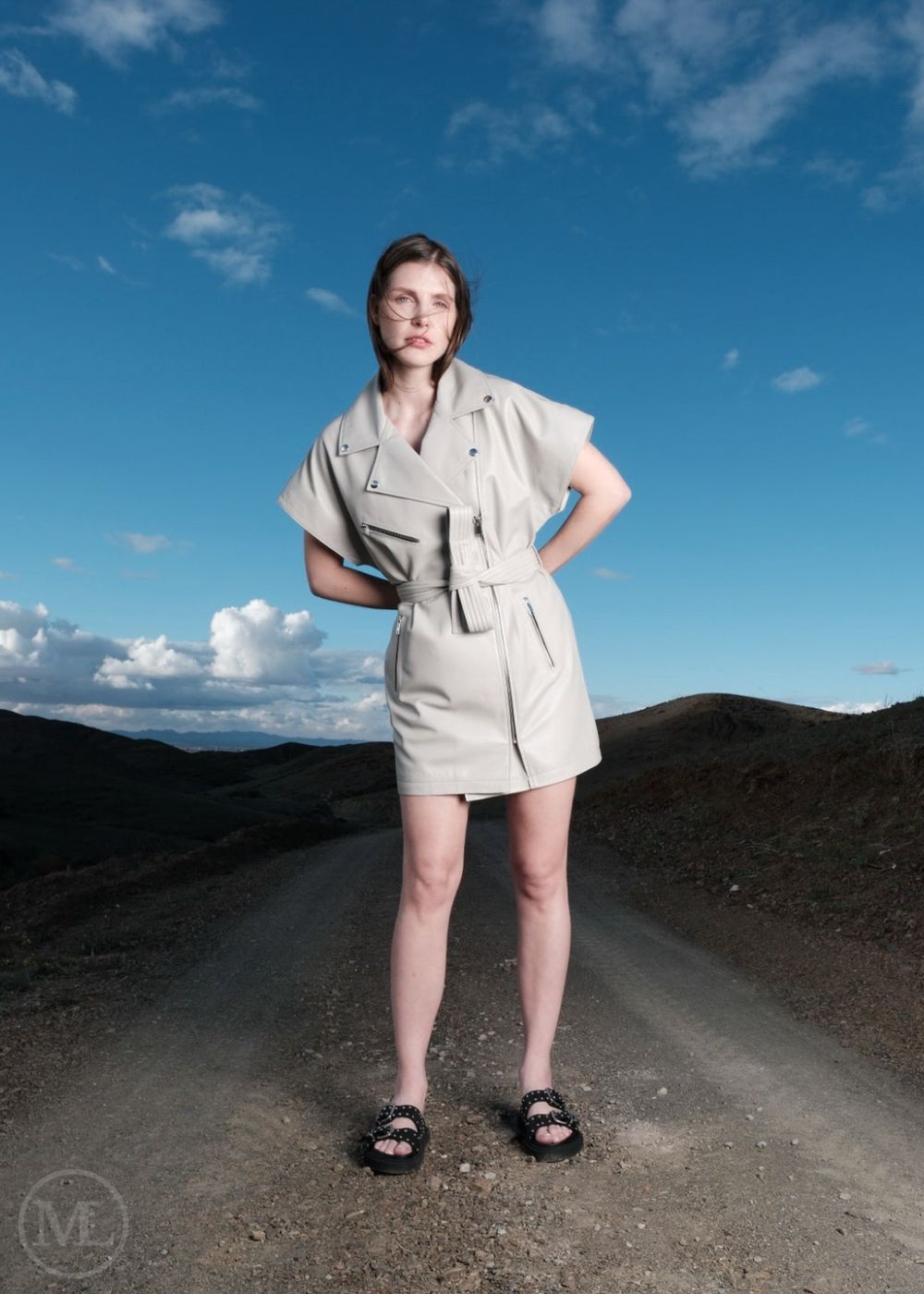 Woman in a ivory leather biker style dress standing on a dirt road with a blue sky and clouds in the background