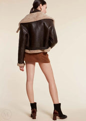 Woman wearing a dark brown aviator jacket with fur lining, brown skirt, and black boots on a beige background.