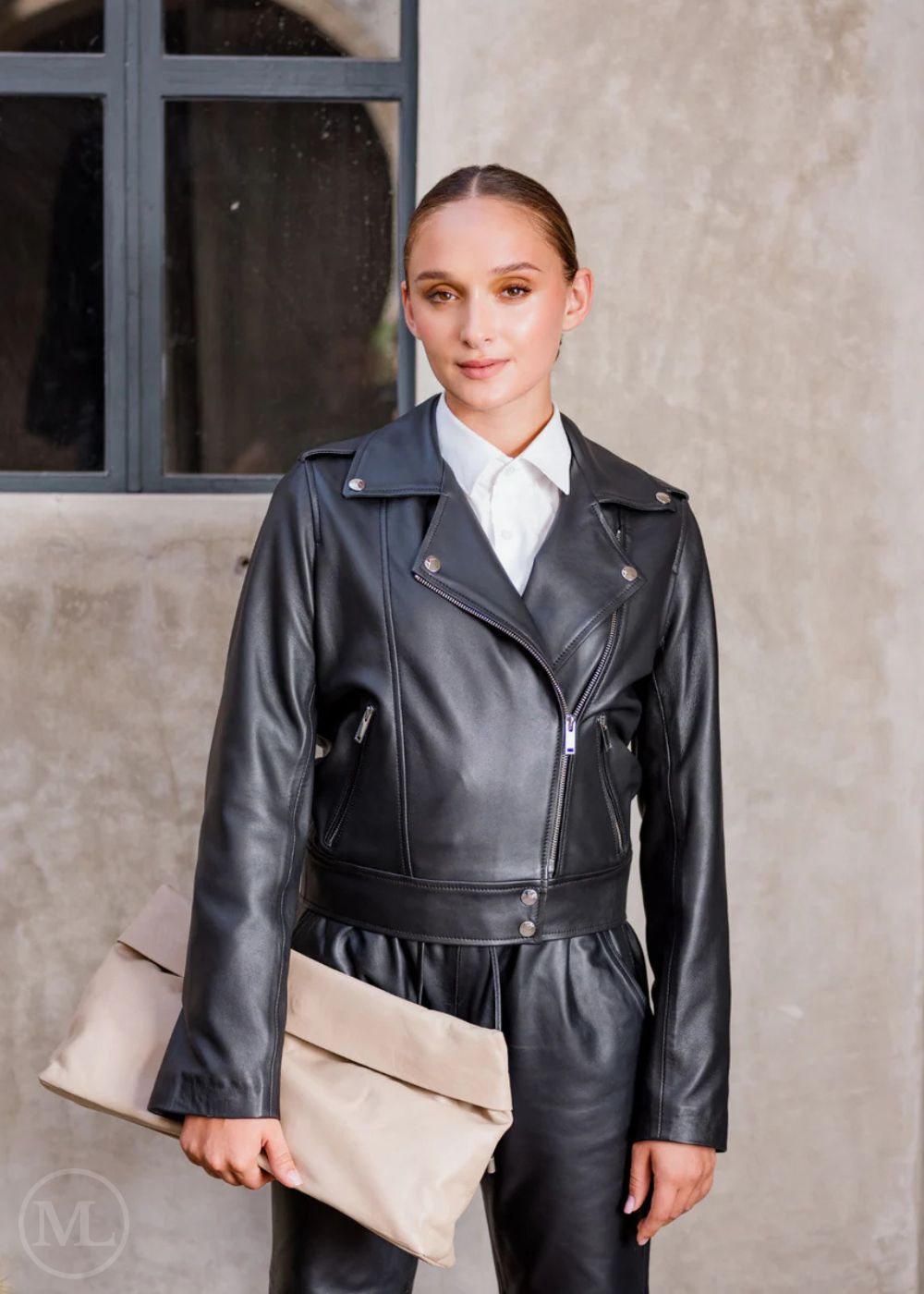 Woman wearing a black leather biker jacket and holding a beige clutch against a neutral background