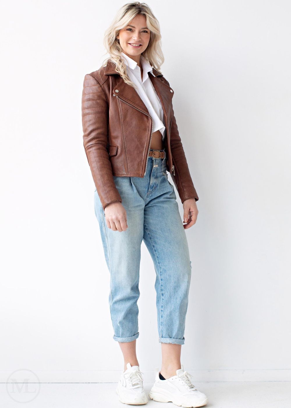 Woman wearing a cognac brown leather jacket, white shirt, light blue jeans, and white sneakers on a white background