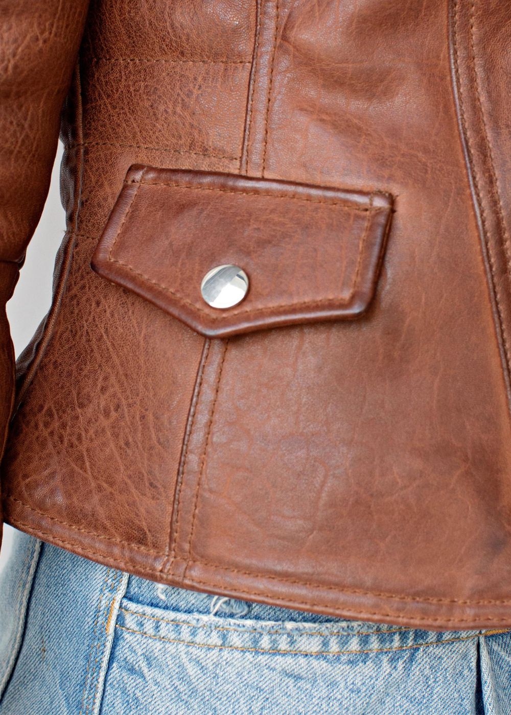 Close-up of a cognac brown leather jacket with a button, worn over blue jeans.