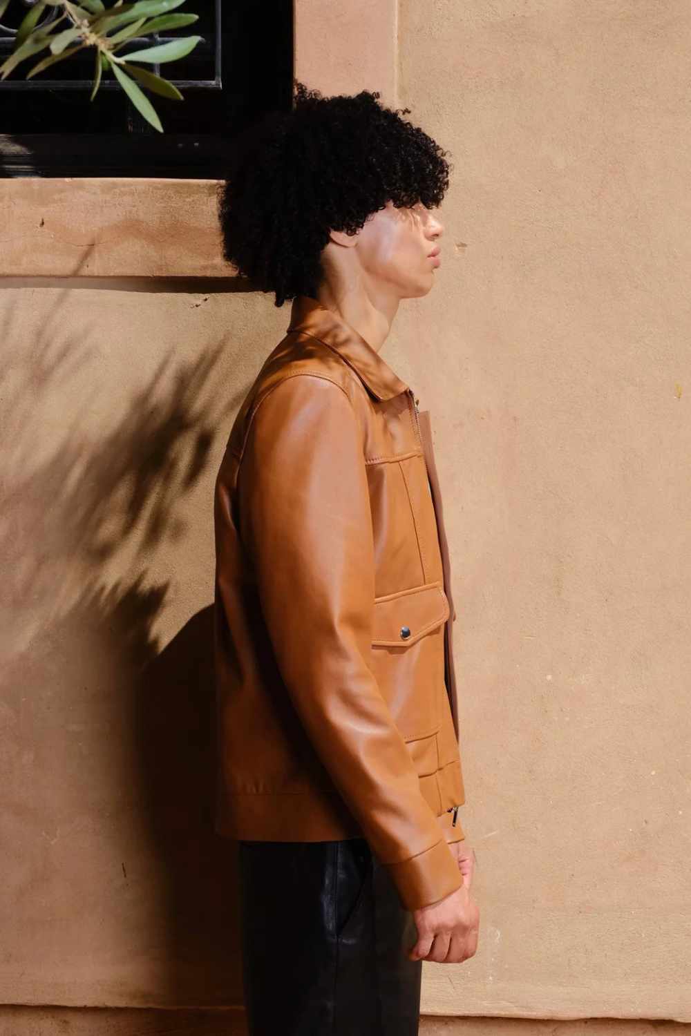 a young boy with curly hair giving side a pose while wearing a Designer men’s camel color leather trucker jacket with flap chest pockets, and welt side pockets,
