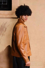 a young boy with curly hair giving a pose while seeing on floor wearing a Designer men’s camel color leather trucker jacket with flap chest pockets, and welt side pockets,
