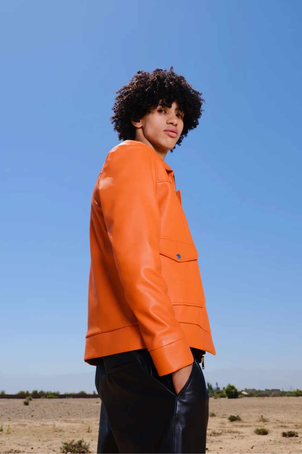 a young boy with curly hair standing in a side pose wearing a Designer men’s orange leather trucker jacket with flap chest pockets, and welt side pockets,
