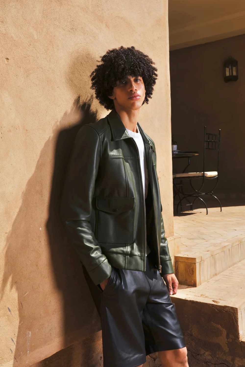 a young boy standing against beige wall. wearing a Designer men’s khaki leather trucker jacket with flap chest pockets, and welt side pockets, close shot