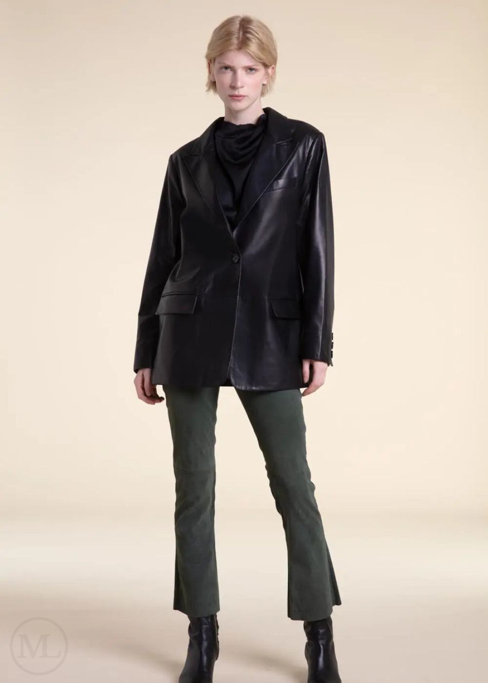 Woman wearing a black leather blazer jacket, black top, green pants, and black boots on a beige background