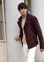 Man wearing a dark brown suede jacket, beige turtleneck, and white pants against a light gray wall.
